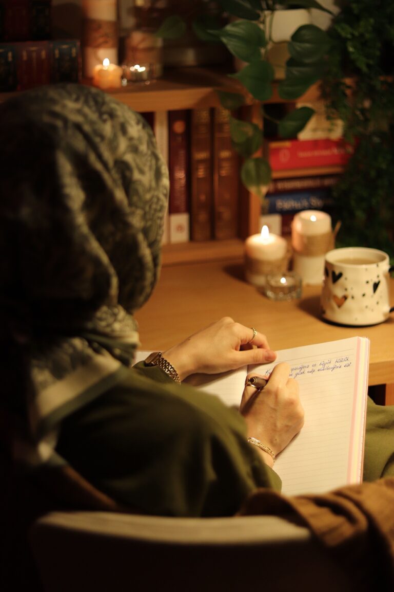 Woman in hijab writing in a journal at a cozy, candle-lit desk setting.